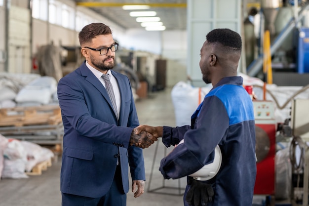 Młody elegancki biznesmen w garniturze i okularach ściska rękę afrykańskiego pracownika dużego zakładu przemysłowego po zawarciu umowy i podpisaniu umowy