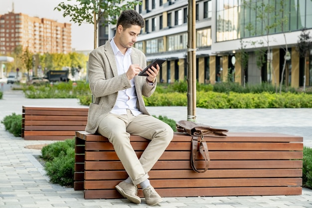 Młody elegancki spokojny biznesmen w stroju formalnym siedzi na drewnianej ławce w parku i przewija wiadomości online w środowisku miejskim