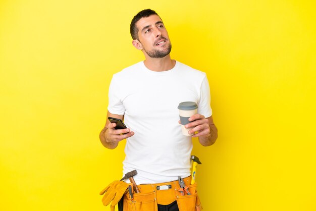 Młody elektryk kaukaski mężczyzna na żółtym tle trzymający kawę na wynos i telefon komórkowy, myśląc o czymś