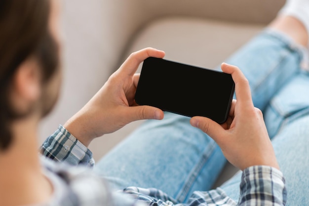 Młody Europejczyk ogląda gry wideo w grze na smartfonie z pustym ekranem na kanapie w salonie