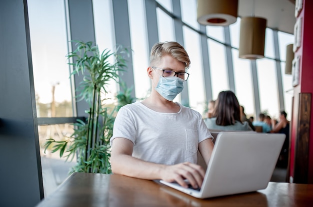 Młody facet w masce medycznej pracuje na laptopie w kawiarni