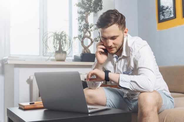 Młody freelancer, patrząc na zegarek i mówiąc na telefon podczas pracy z domu