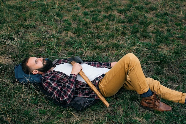 Młody hipster mężczyzna leżący na zielonej trawie w lesie z siekierą w rękach Koncepcja rekreacji na świeżym powietrzu