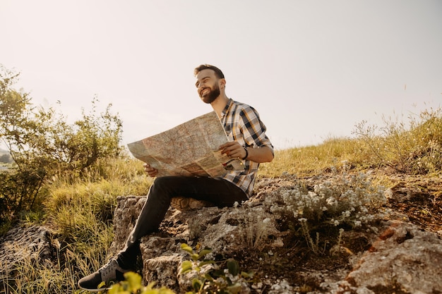 Młody hipster mężczyzna uśmiechnięty, siedzący na kamieniu, trzymając mapę.