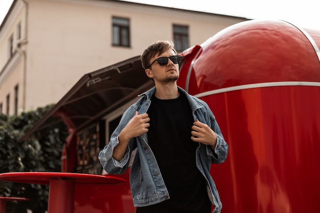 Młody hipster miejski zdejmuje modną kurtkę dżinsową. Model atrakcyjny facet w okulary pozuje na tle czerwonej furgonetki z jedzeniem w mieście w słoneczny wiosenny dzień. Modna odzież męska.