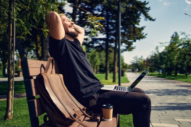 Młody hipster milenijny siedzi na ławce w parku z laptopem i cieszy się wolnym czasem