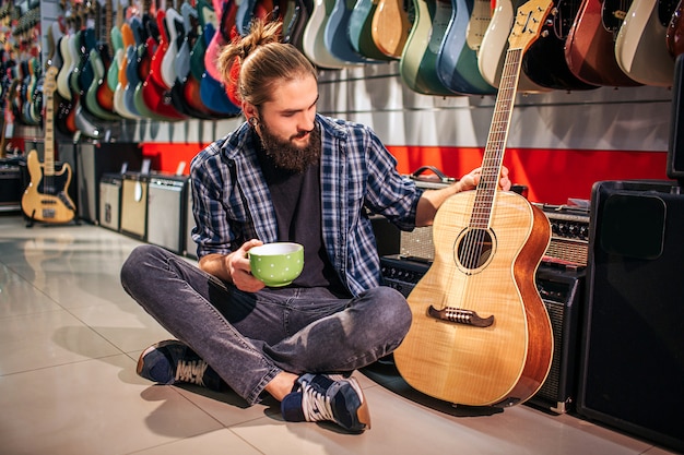 Młody hipster odpoczywa, siedząc na podłodze. Trzyma kubek w ręku. Kolejny dotyka pobliskiej gitary akustycznej. Młody człowiek patrzy na instrument.