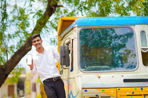 Młody indyjski biznesmen ze swoją ciężarówką lub ciężarówką do przodu i pokazując smartphone.