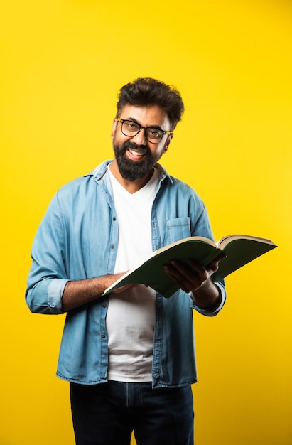 Młody indyjski szczęśliwy mężczyzna noszący okulary, czytający książkę i uśmiechający się na żółto