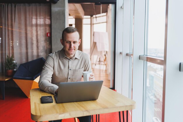 Młody inteligentny człowiek siedzi w biurze z filiżanką kawy i pracuje nad projektem związanym z publiczną technologią cybernetyczną Biznesmeni z laptopem próbują dotrzymać terminów w marketingu zewnętrznym