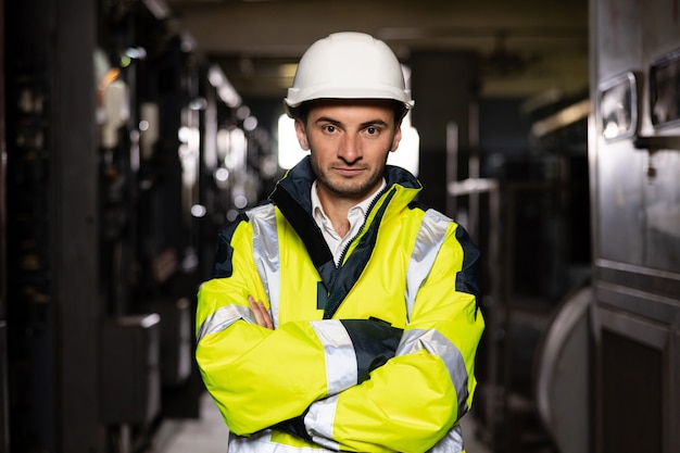 Młody inżynier fabryki lub pracownik noszący kamizelkę odblaskową i kask krzyżujący ramiona w pomieszczeniu elektrycznym