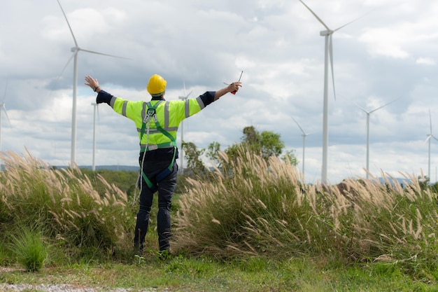 Młody inżynier pracujący na polu turbin wiatrowych nosi kamizelkę bezpieczeństwa, podnosząc obie ręce, aby się zrelaksować po zakończeniu misji kontrolnej turbiny wiatrowej