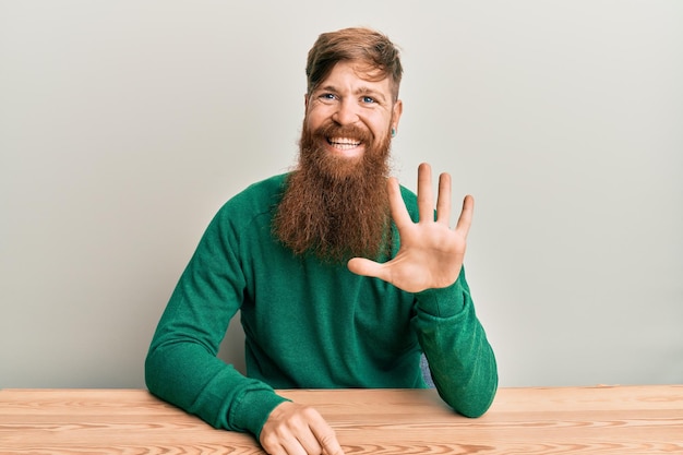 Młody irlandczyk rudy mężczyzna ubrany w zwykłe ubrania, siedzący na stole, pokazujący i wskazujący palcami numer pięć, uśmiechając się pewny siebie i szczęśliwy.
