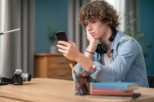 Młody Kanadyjczyk używający smartfona, leżąc przy biurku w swoim salonie, nudny męski telefon