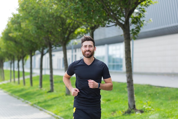 Młody kaukaski brodaty człowiek szczęśliwy biegacz jogging ulicą na tle miejskim parku miejskiego. Poranny jogging. Aktywny zdrowy styl życia fitness. Trening sportowy na świeżym powietrzu. Trening sportowca bieganie ćwiczenie