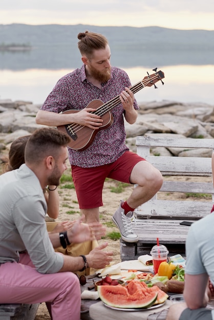 Młody kaukaski brodaty mężczyzna gra na gitarze podczas spędzania czasu z przyjaciółmi poza miastem w pobliżu jeziora w piękny letni wieczór