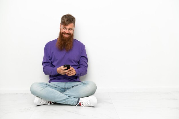 Młody kaukaski czerwonawy mężczyzna siedzący na podłodze na białym tle wysyłający wiadomość za pomocą telefonu komórkowego