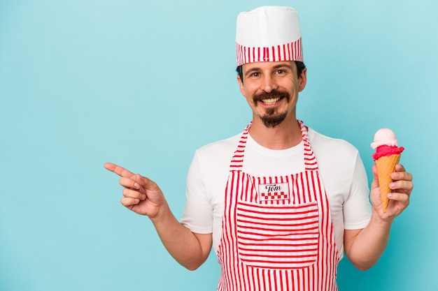 Młody kaukaski lodziarz trzymający lody na białym tle na niebieskim tle, uśmiechając się i wskazując na bok, pokazując coś w pustej przestrzeni.