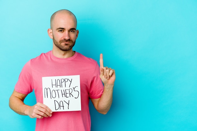Młody kaukaski łysy mężczyzna trzyma afisz szczęśliwy dzień matki na białym tle na niebieskiej ścianie pokazuje numer jeden z palcem.