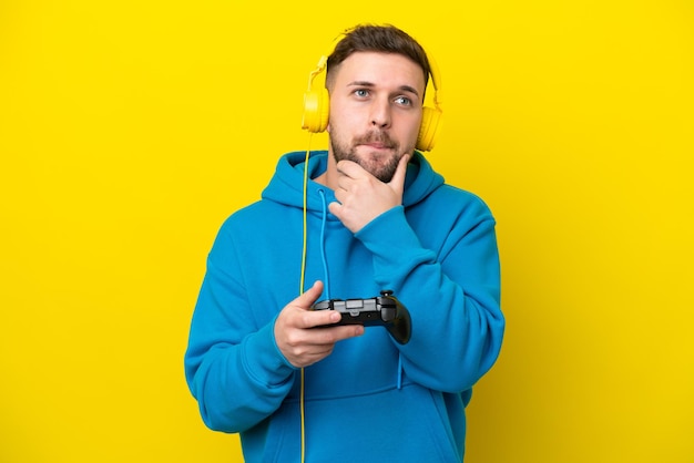 Zdjęcie młody kaukaski mężczyzna bawiący się kontrolerem gier wideo na żółtym tle, mający wątpliwości i myślący