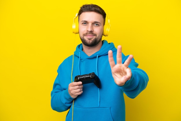 Młody kaukaski mężczyzna bawiący się kontrolerem gier wideo na żółtym tle szczęśliwy i liczący trzy palcami
