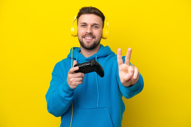 Młody kaukaski mężczyzna bawiący się kontrolerem gier wideo na żółtym tle, uśmiechający się i pokazujący znak zwycięstwa