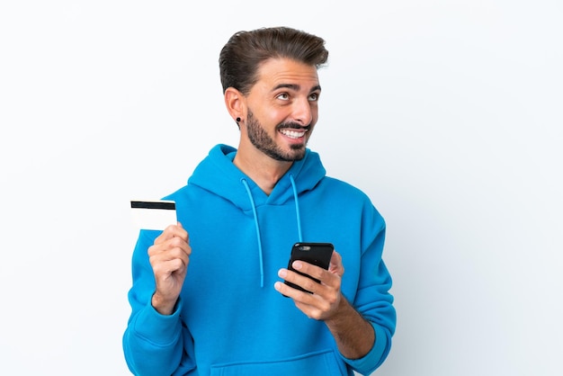 Młody kaukaski mężczyzna na białym tle kupując za pomocą telefonu komórkowego za pomocą karty kredytowej podczas myślenia