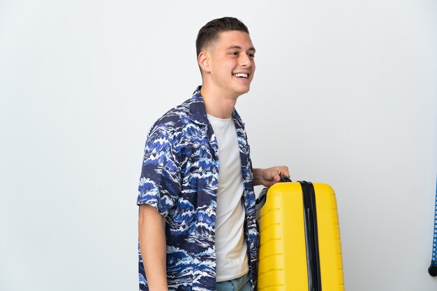 Młody kaukaski mężczyzna na białym tle na białej ścianie w wakacje z walizką podróżną