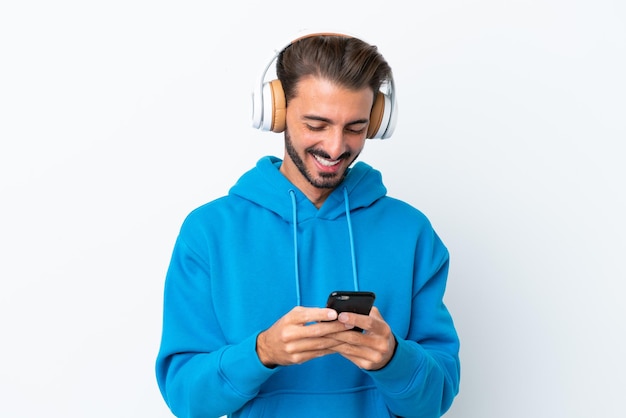 Młody kaukaski mężczyzna na białym tle słuchający muzyki i patrzący na telefon komórkowy