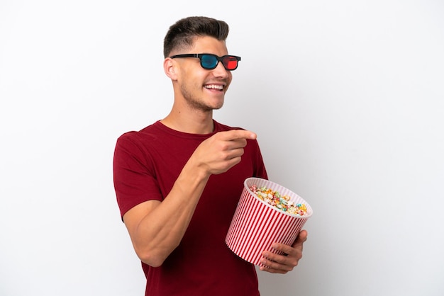 Młody kaukaski mężczyzna na białym tle w okularach 3d i trzymający duże wiadro popcornów, wskazując przód