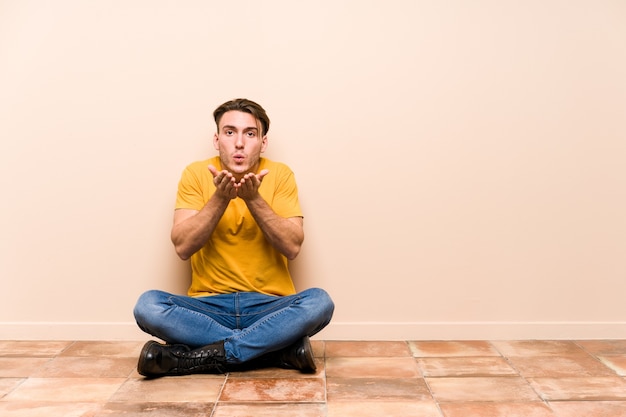 Młody kaukaski mężczyzna siedzi na podłodze na białym tle składane usta i trzymając dłonie, aby wysłać pocałunek.