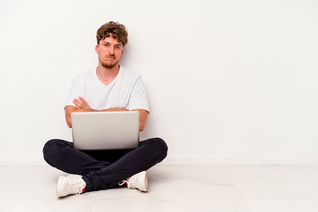 Młody kaukaski mężczyzna siedzi na podłodze trzymając laptopa na białym tle niezadowolony patrząc w kamerę z sarkastycznym wyrazem.