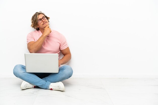 Młody kaukaski mężczyzna siedzi na podłodze z laptopem na białym tle i patrząc w górę