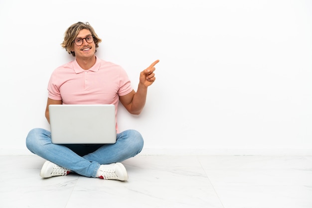 Młody kaukaski mężczyzna siedzi na podłodze z laptopem na białym tle na biały palcem wskazującym na bok