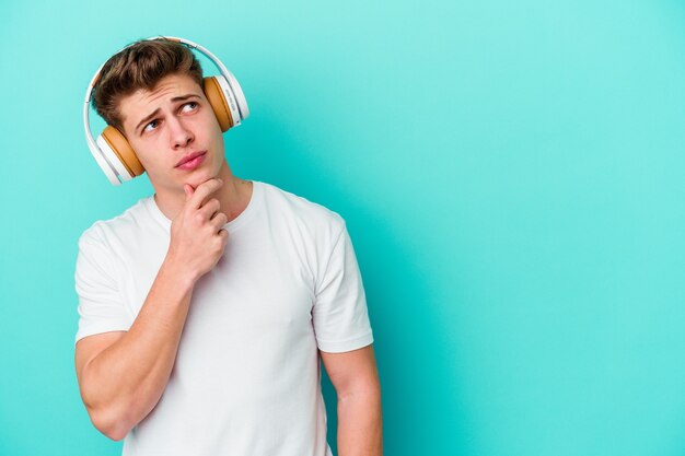 Młody kaukaski mężczyzna słucha muzyki w słuchawkach na niebiesko, patrząc z boku z wątpliwym i sceptycznym wyrazem.