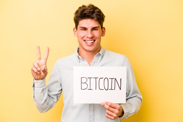 Młody kaukaski mężczyzna trzyma afisz bitcoin na białym tle na żółtym tle pokazujący numer dwa palcami