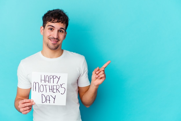 Młody kaukaski mężczyzna trzyma afisz szczęśliwy dzień matki na białym tle, uśmiechając się i wskazując na bok, pokazując coś w pustej przestrzeni.