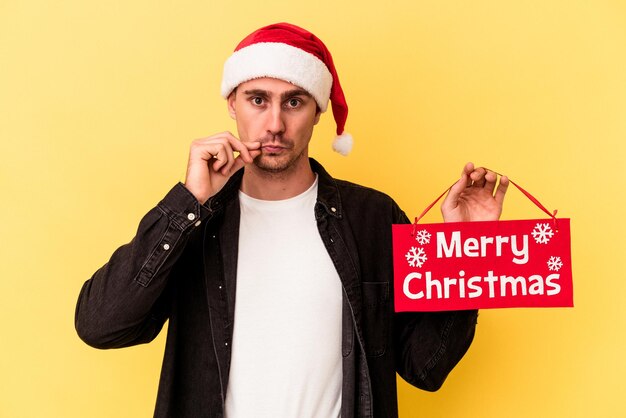 Młody kaukaski mężczyzna trzyma afisz Wesołych Świąt na białym tle na żółtym tle z palcami na ustach, zachowując tajemnicę.