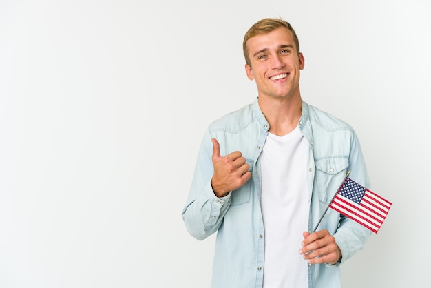 Młody kaukaski mężczyzna trzyma amerykańską flagę na białym tle na białej ścianie, uśmiechając się i podnosząc kciuk do góry