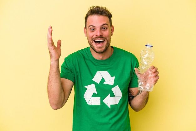 Młody kaukaski mężczyzna trzyma butelkę plastiku do recyklingu na białym tle na żółtym tle otrzymując miłą niespodziankę, podekscytowany i podnosząc ręce.