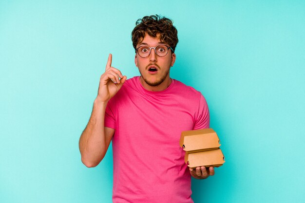 Młody kaukaski mężczyzna trzyma dwa hamburgery na białym tle na niebieskim tle o pomysł, koncepcja inspiracji.
