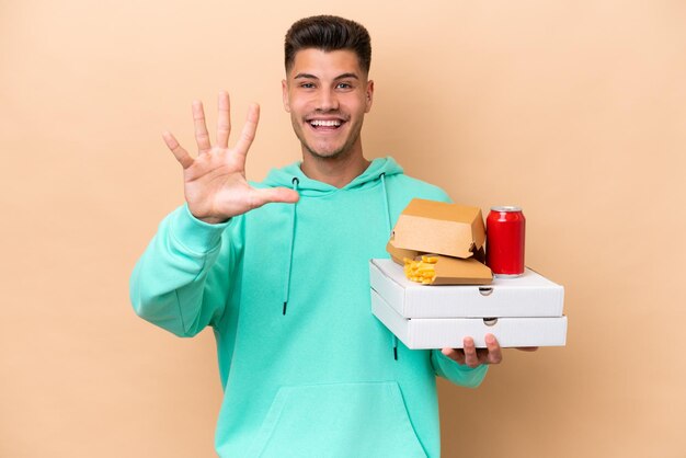 Młody kaukaski mężczyzna trzyma fast food na białym tle na beżowym tle, licząc pięć palcami