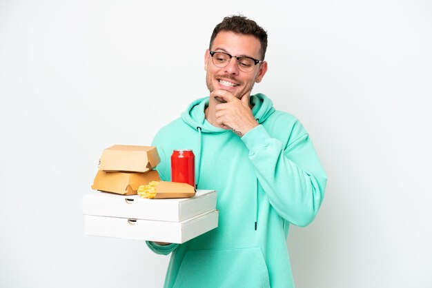 Młody kaukaski mężczyzna trzyma fast food na białym tle, patrząc z boku i uśmiechając się