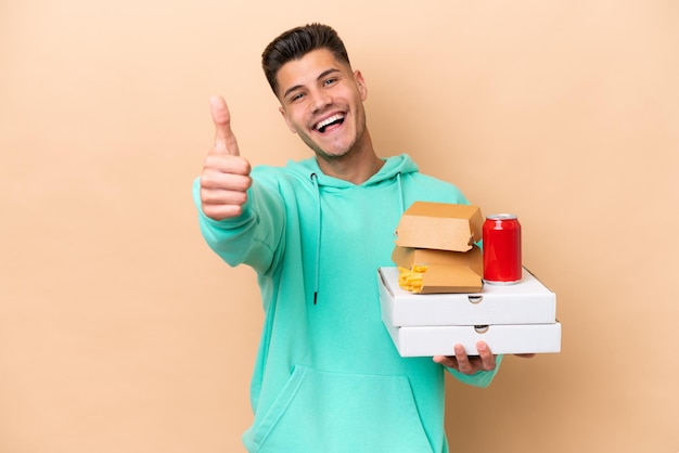 Młody kaukaski mężczyzna trzyma fast food odizolowany na beżowym tle z kciukami do góry, ponieważ stało się coś dobrego