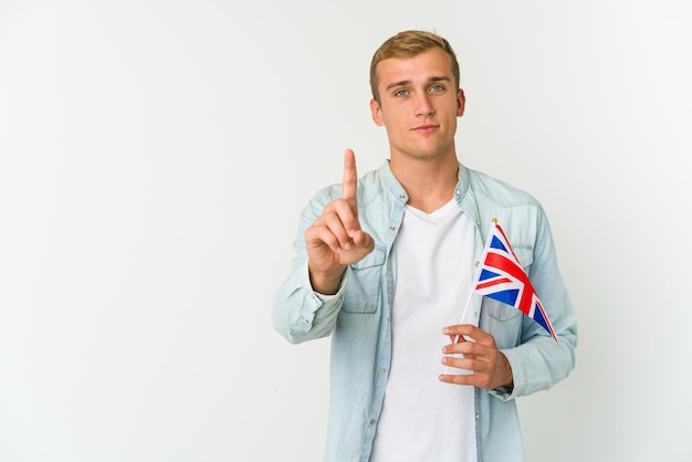Młody Kaukaski Mężczyzna Trzyma Flagę Wielkiej Brytanii Na Białym Tle Na Białej Przestrzeni Pokazuje Numer Jeden Z Palcem.