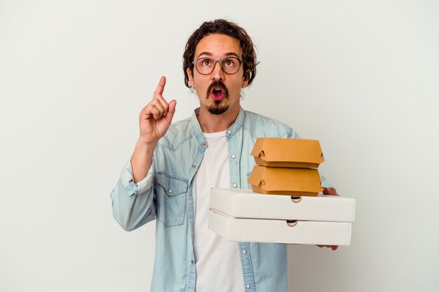 Młody kaukaski mężczyzna trzyma hamburgera i pizze na białym tle na białej ścianie, wskazując do góry z otwartymi ustami.