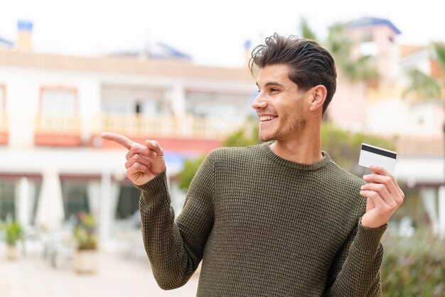 Młody kaukaski mężczyzna trzyma kartę kredytową na zewnątrz, wskazując na bok, aby zaprezentować produkt