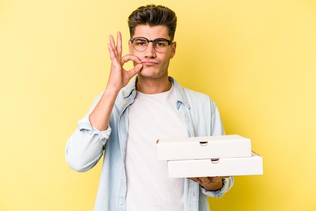 Młody kaukaski mężczyzna trzyma pizze na białym tle na żółtym tle z palcami na ustach zachowując tajemnicę.