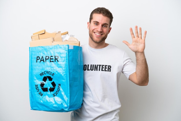 Młody kaukaski mężczyzna trzyma torbę do recyklingu pełną papieru do recyklingu na białym tle na białym tle pozdrawiając ręką z szczęśliwym wyrazem twarzy