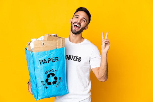 Młody kaukaski mężczyzna trzyma torbę do recyklingu pełną papieru do recyklingu na białym tle, uśmiechając się i pokazując znak zwycięstwa